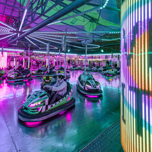 Der Autoscooter ist bunt beleuchtet. Im Vordergrund sieht man eine helle Lichtersäule und im Hintergrund begeisterte Besucher, die auf der Fläche mit den Chaisen fahren